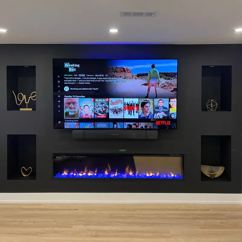 A modern living room with a wall-mounted TV displaying Netflix, a sleek black electric fireplace below, and decorative shelves on both sides holding various art pieces.