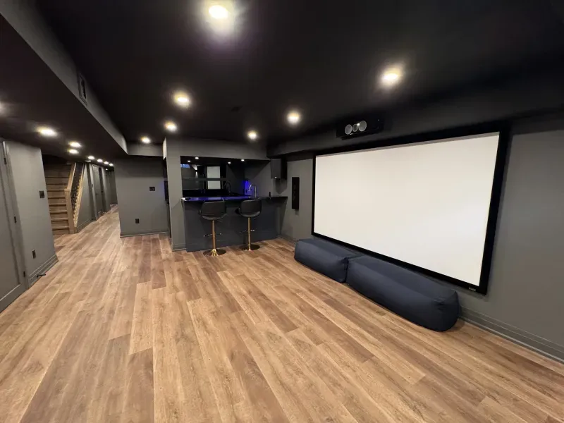Modern home theater room with wood flooring, a large projector screen, two floor cushions, a small bar area with two stools, and recessed ceiling lights. A staircase is visible in the background.
