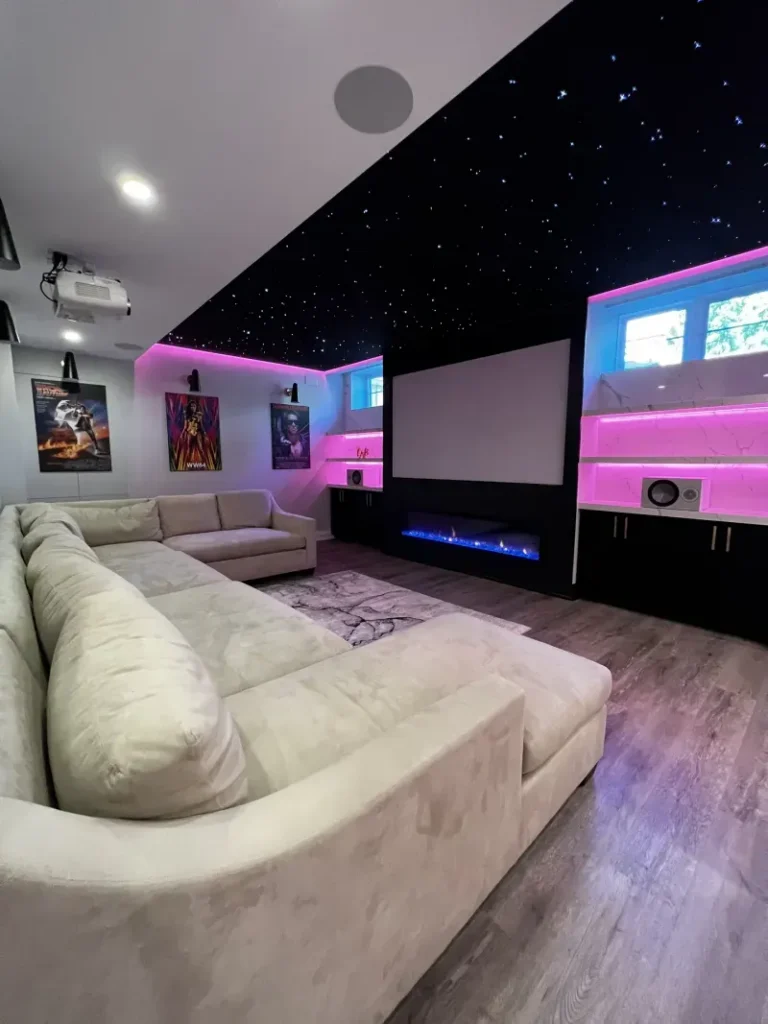 Modern home theater with a large sectional sofa, wall art, a big projector screen, pink and blue LED lights, a black starry ceiling, and wood flooring.