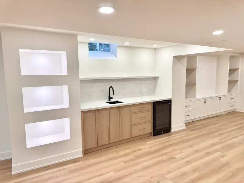 Modern basement kitchenette with light wood cabinets, black faucet, small sink, under-counter fridge, open shelving, and built-in white shelving units. Recessed ceiling lights and wood flooring. Small window above counter.
