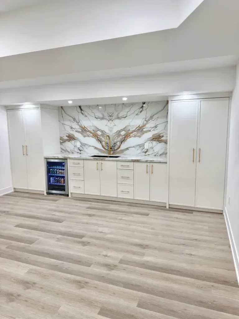 Modern kitchenette with white cabinets, a marble backsplash, a small sink, and a mini fridge stocked with drinks. The room has light wood flooring and recessed ceiling lights above the counter area.