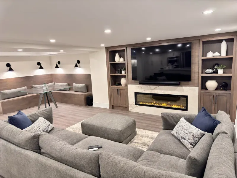 Modern living room with a large sectional sofa, ottoman, wall-mounted TV, built-in shelves, electric fireplace, and a nook with bench seating and a glass table. Neutral colors and soft lighting create a cozy atmosphere.