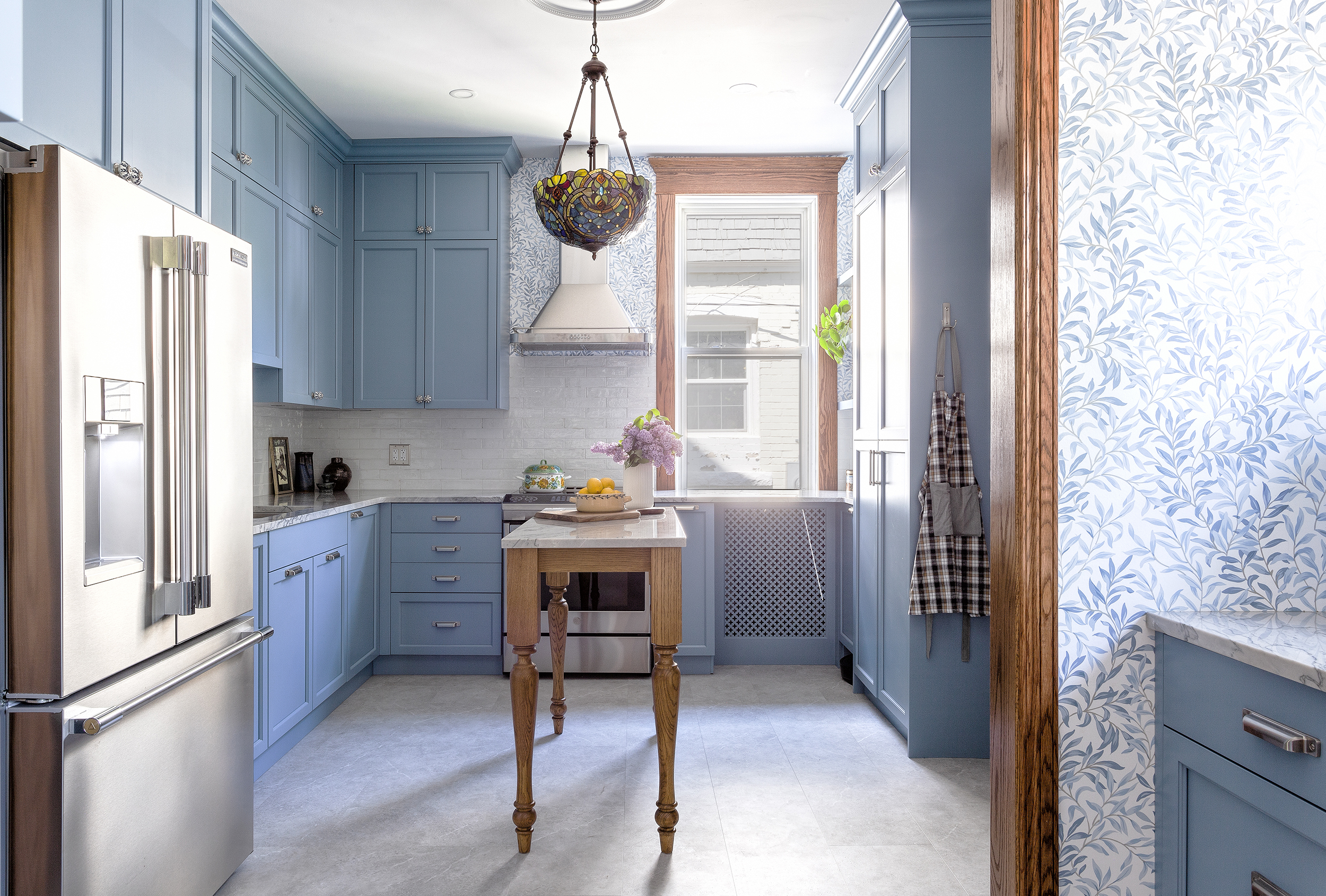 A bright kitchen with blue cabinets, a wooden island, stainless steel appliances, a floral chandelier, and a window letting in natural light. Flowers and fruit decorate the island.