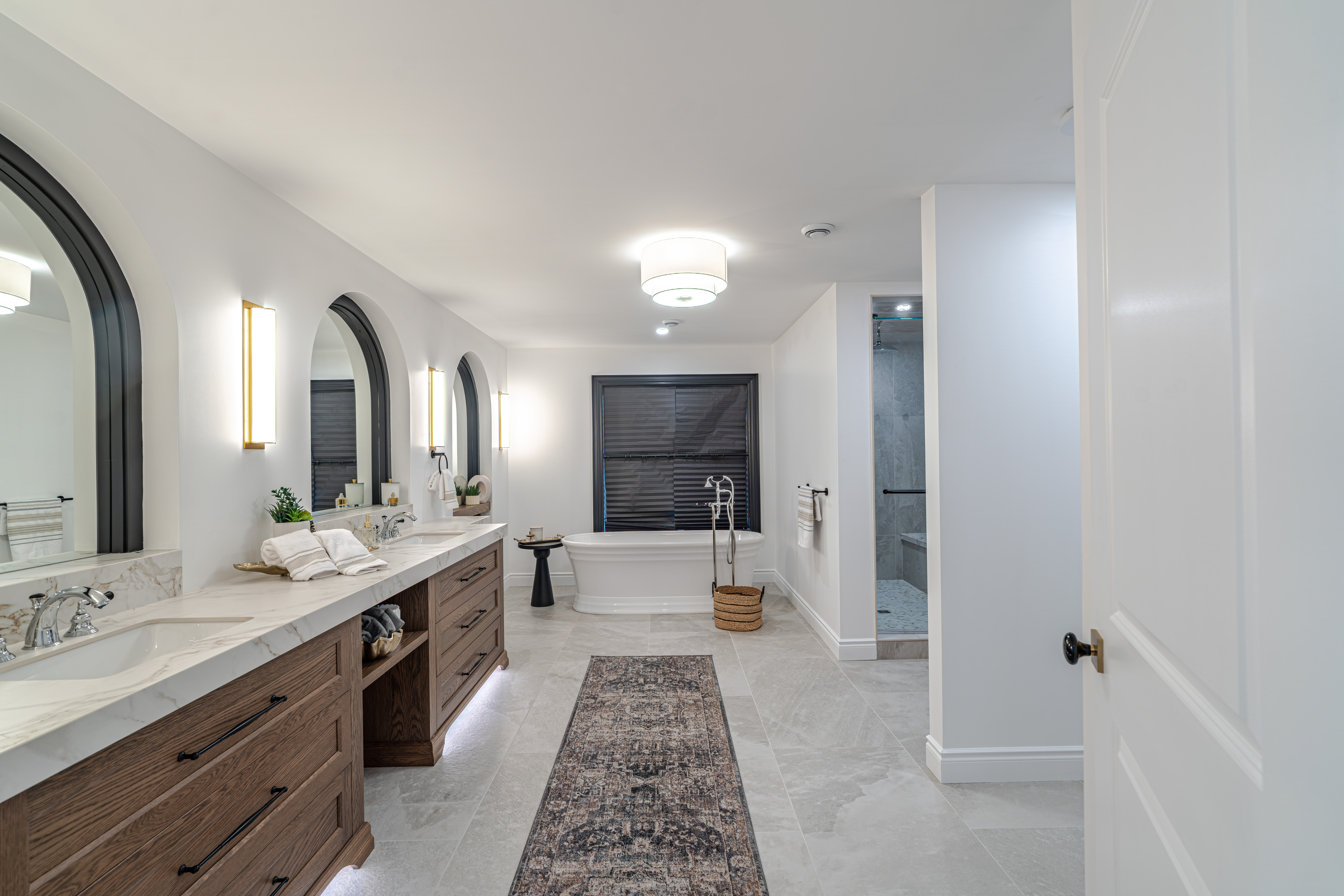 Modern bathroom with double sinks, wooden cabinets, a freestanding tub by a window with blinds, a walk-in shower, and a decorative rug on a light tiled floor under ceiling lights.