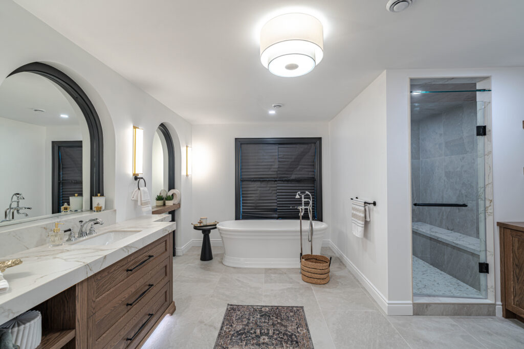 Modern bathroom with double vanity, freestanding bathtub, glass shower, and neutral decor. Marble countertops, wooden cabinets, and a large window with dark blinds are featured. Contemporary lighting brightens the space.
