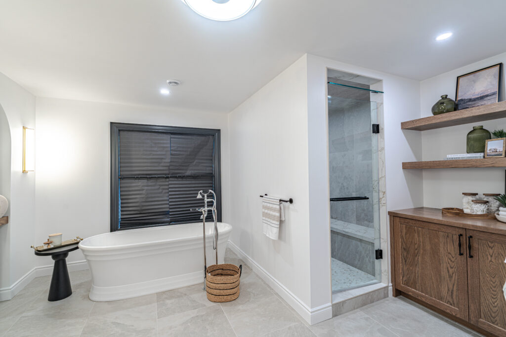 Modern bathroom with a freestanding tub, glass shower, wooden shelves with decor, a wicker basket, and a cabinet. The space has neutral colors and soft lighting.