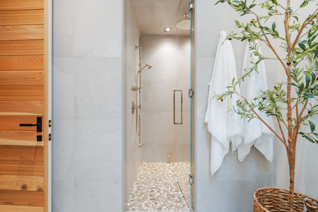 A modern bathroom with a stone floor walk-in shower, glass door, wooden door on the left, two white towels hanging on hooks, and a leafy plant in a woven basket.