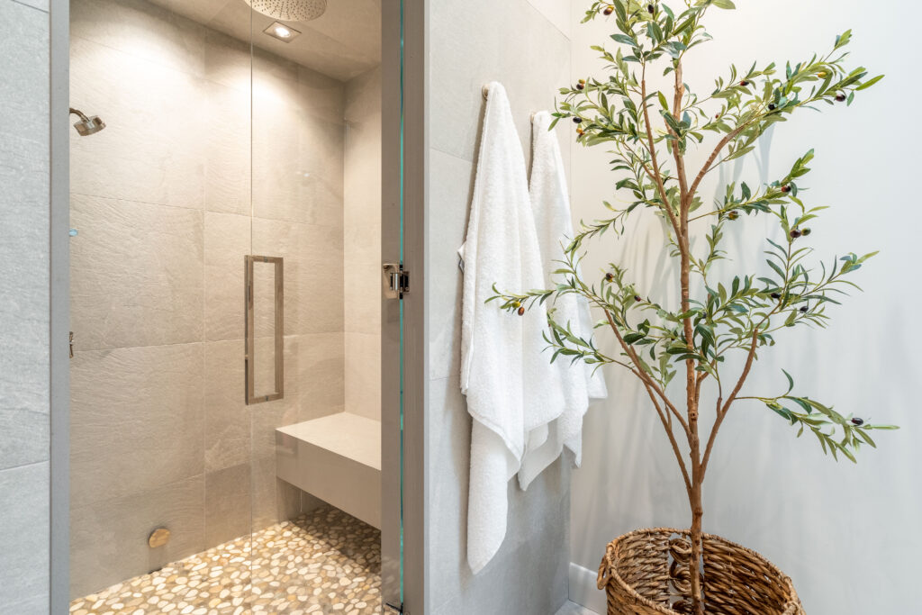 Modern bathroom with a glass-enclosed shower, stone tile floor, two white towels hanging on the wall, and a potted plant with green leaves in a woven basket nearby.