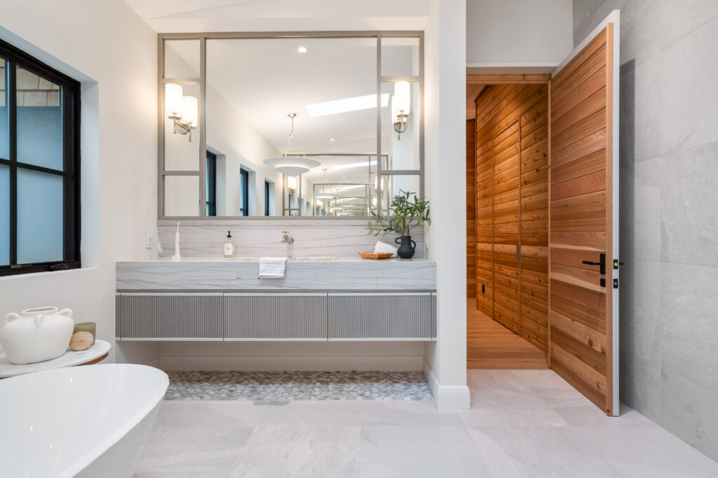 Modern bathroom with a white bathtub, floating vanity, large mirrors, and a wooden door leading to a sauna. There are light fixtures, a plant, and natural light from windows. Surfaces are clean and bright.