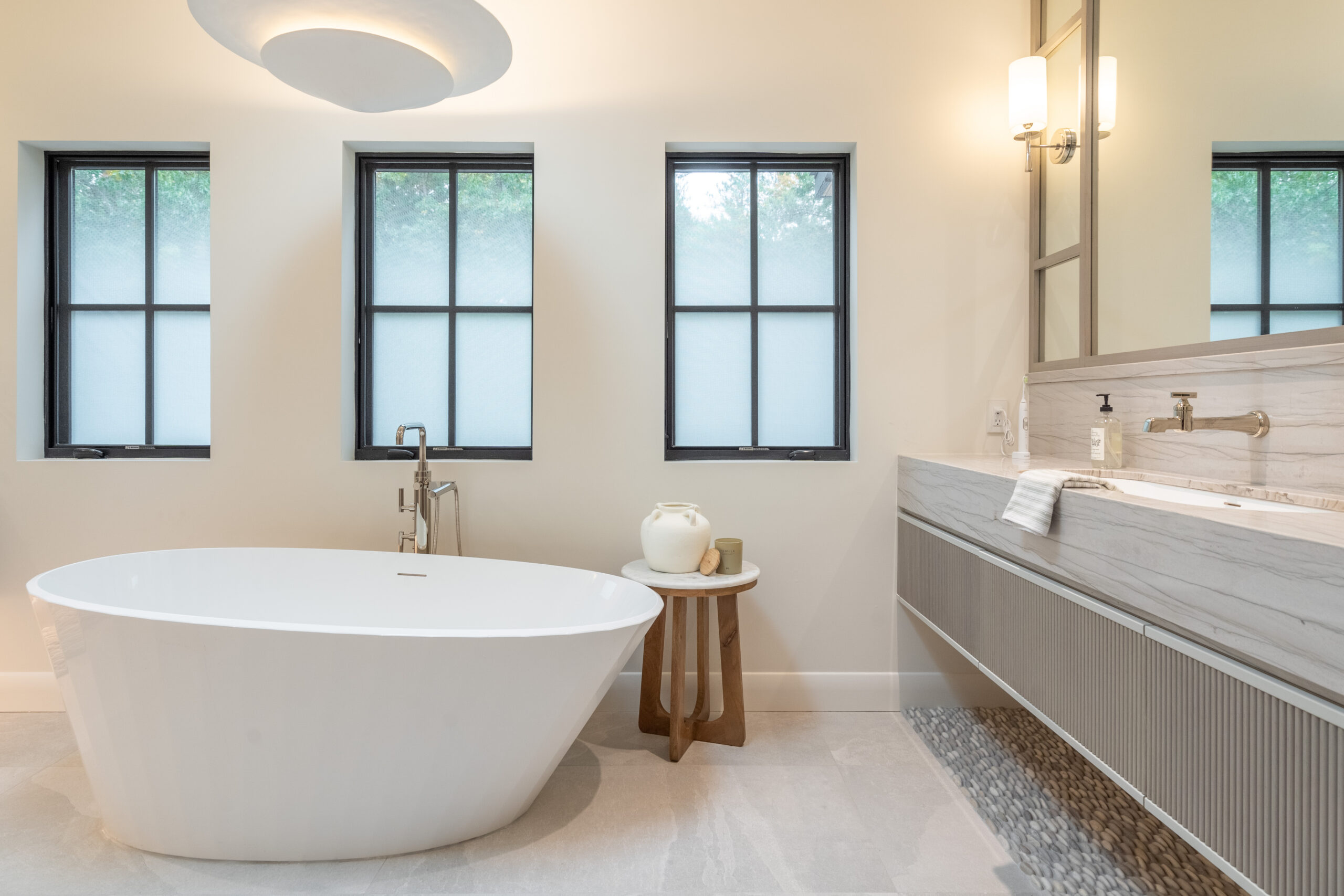 Modern bathroom with a white freestanding tub, small wooden side table, double sink vanity, and three frosted windows. Neutral tones and minimalist decor create a clean, serene atmosphere.