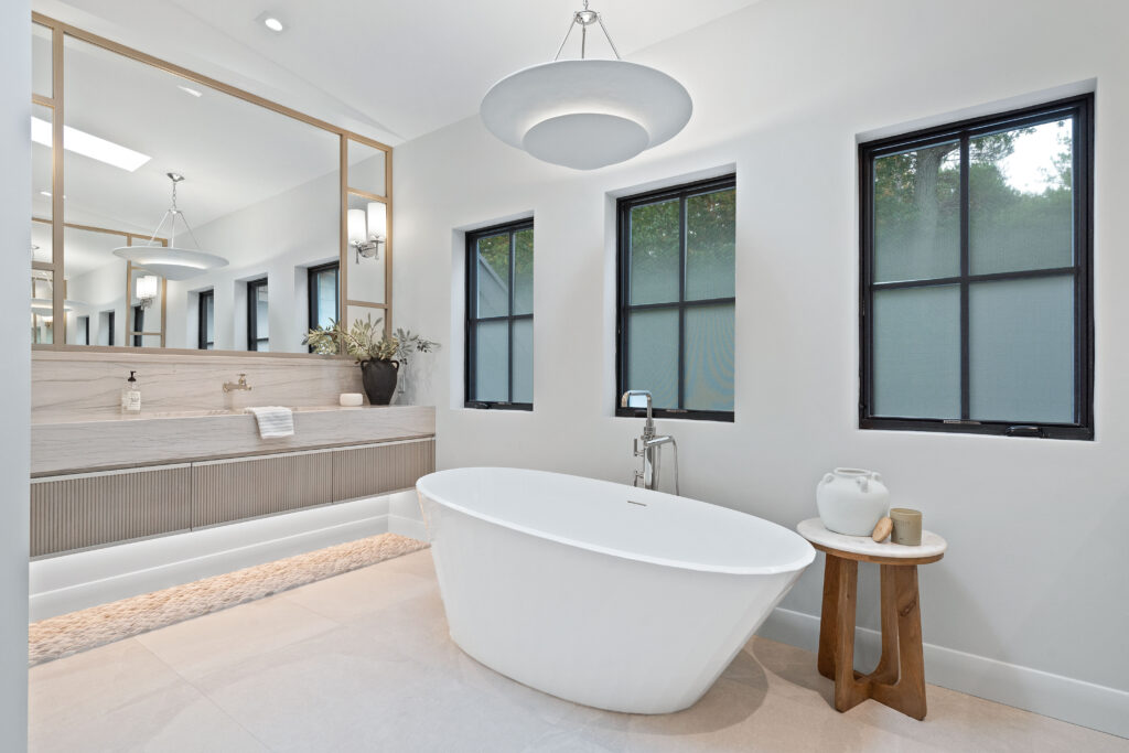 Modern bathroom with a white freestanding tub, light wood vanity, large mirrors, two windows with black frames, and a small wooden side table with decor accents. Neutral tones and minimalistic decor create a serene atmosphere.
