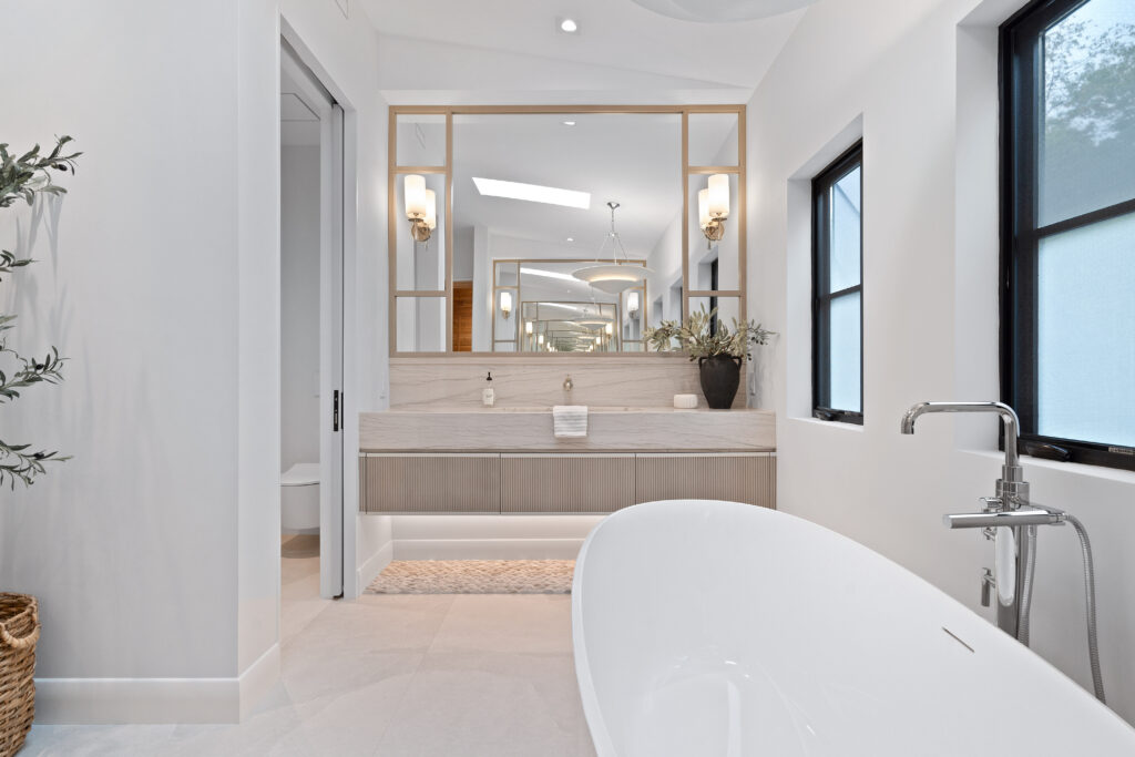 Modern bathroom with a white freestanding bathtub, double sinks, large mirrors, and wall sconces. Natural light comes through two frosted windows. A plant in a woven basket decorates the corner.