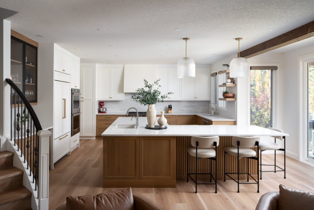 Modern kitchen with white cabinets, wood accents, marble countertops, three stools at an island, pendant lights, open shelves, and large windows bringing in natural light.