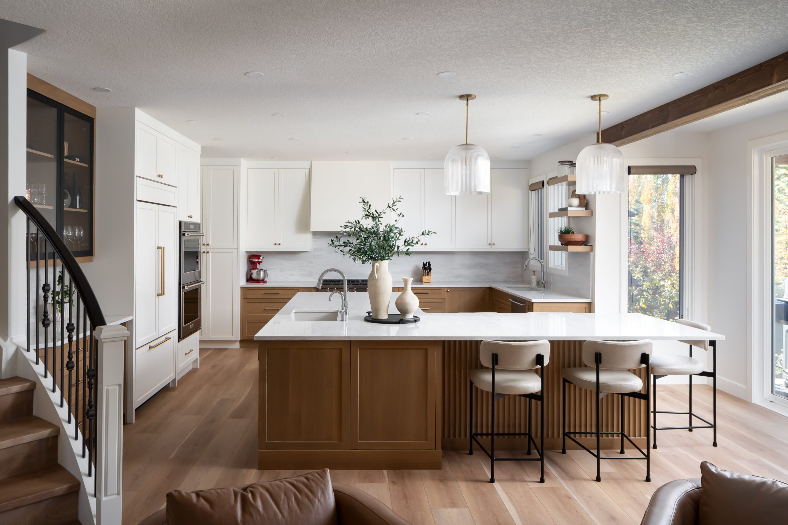 Modern kitchen with white and wood cabinets, marble countertops, three barstools, pendant lights, a vase with greenery on the island, and large windows letting in natural light.