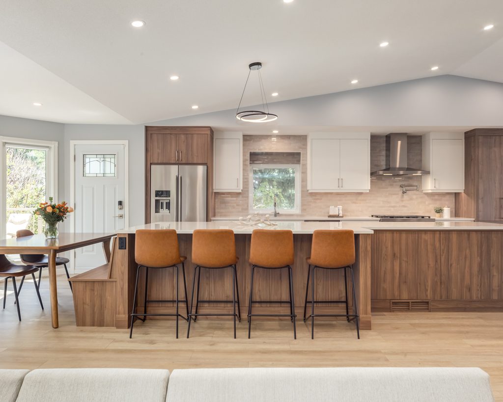 Modern kitchen with wood cabinets, white countertops, four brown barstools at a large island, stainless steel appliances, pendant light, and a dining area with a round table and flowers in a vase.