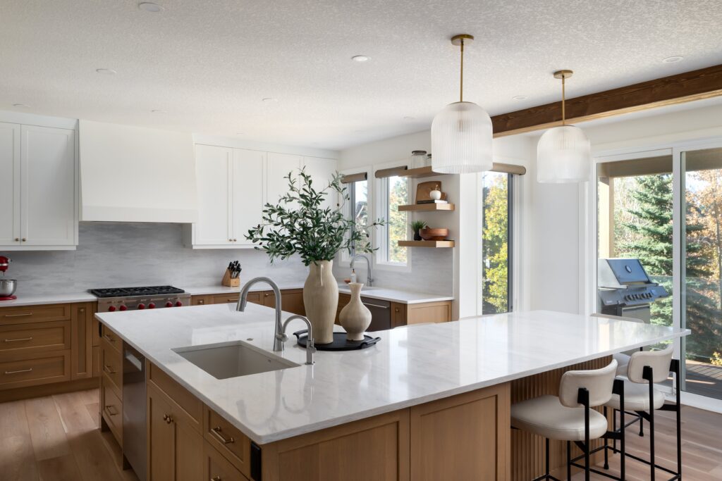 Modern kitchen with a large marble island, wooden cabinets, white upper cabinets, hanging pendant lights, bar stools, a sink, and large windows overlooking greenery. Decorative vases and plants are on the island.