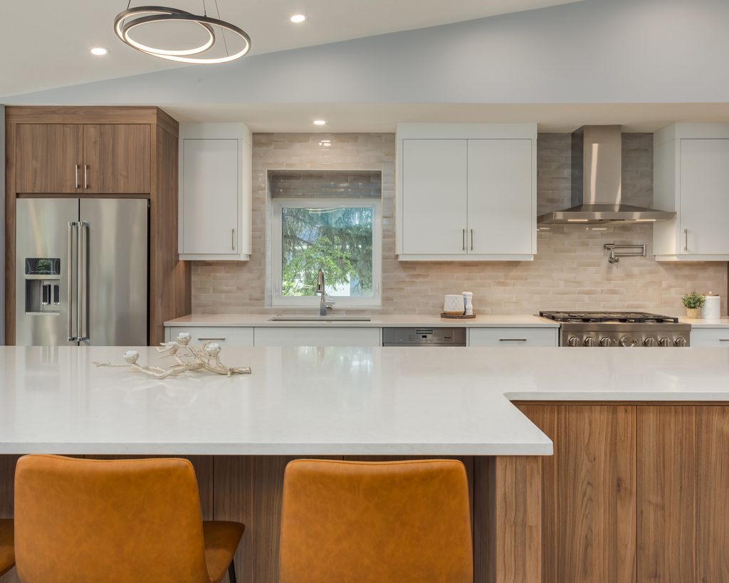 Modern kitchen with wood and white cabinets, stainless steel appliances, a large island with two brown chairs, and a round light fixture above. A window above the sink shows green trees outside.