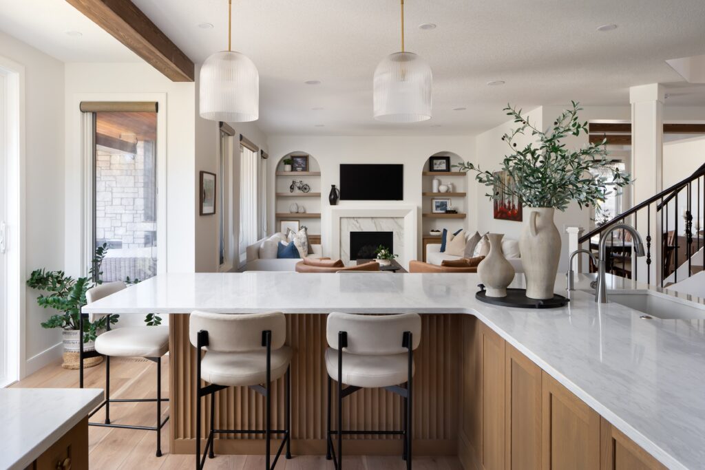 Modern open kitchen with a white marble island, wooden cabinetry, bar stools, and pendant lights, adjoining a cozy living area with a fireplace, TV, and neutral-toned decor.
