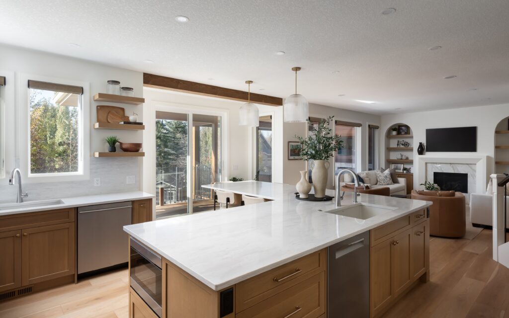 Modern kitchen with wood cabinets, white marble countertops, island sink, open shelves, and pendant lights. Living area with fireplace and TV is visible in the background. Large windows bring in natural light.