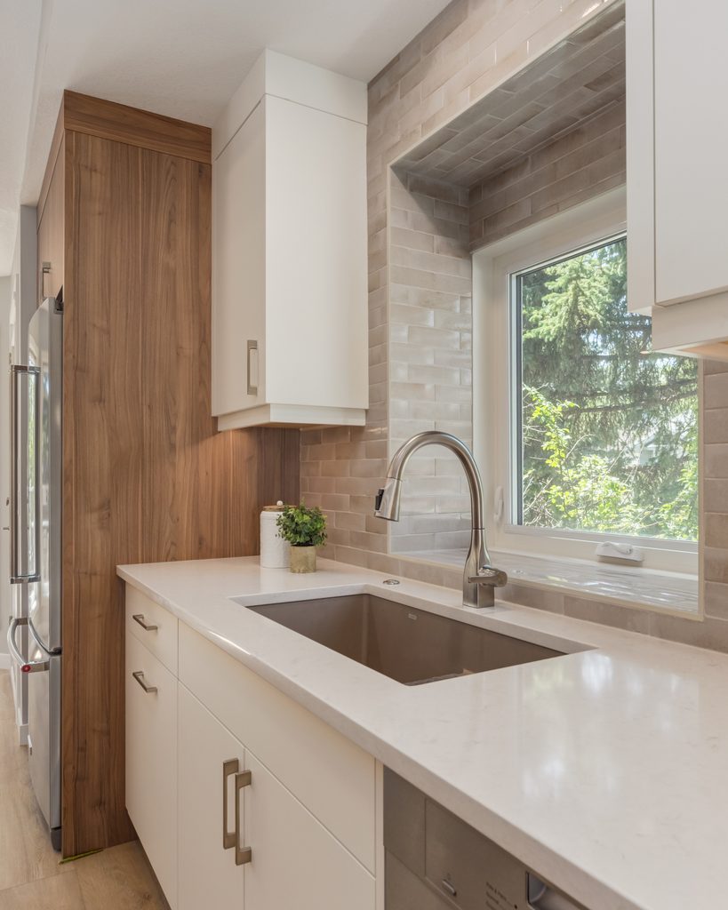 Modern kitchen with white cabinets, a wood accent wall, stainless steel sink, faucet, dishwasher, and a window with a view of green trees outside. Countertop has a small potted plant and a container.