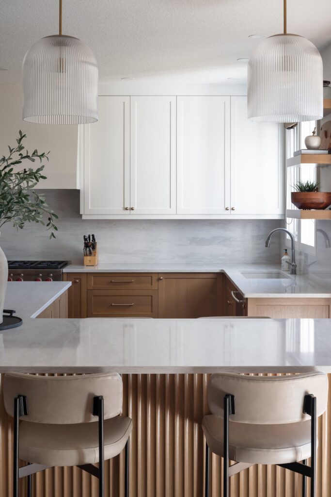 Modern kitchen with white upper cabinets, wood lower cabinets, a marble countertop, two beige barstools, pendant lights, and a plant on the left. The space is bright and neatly organized.