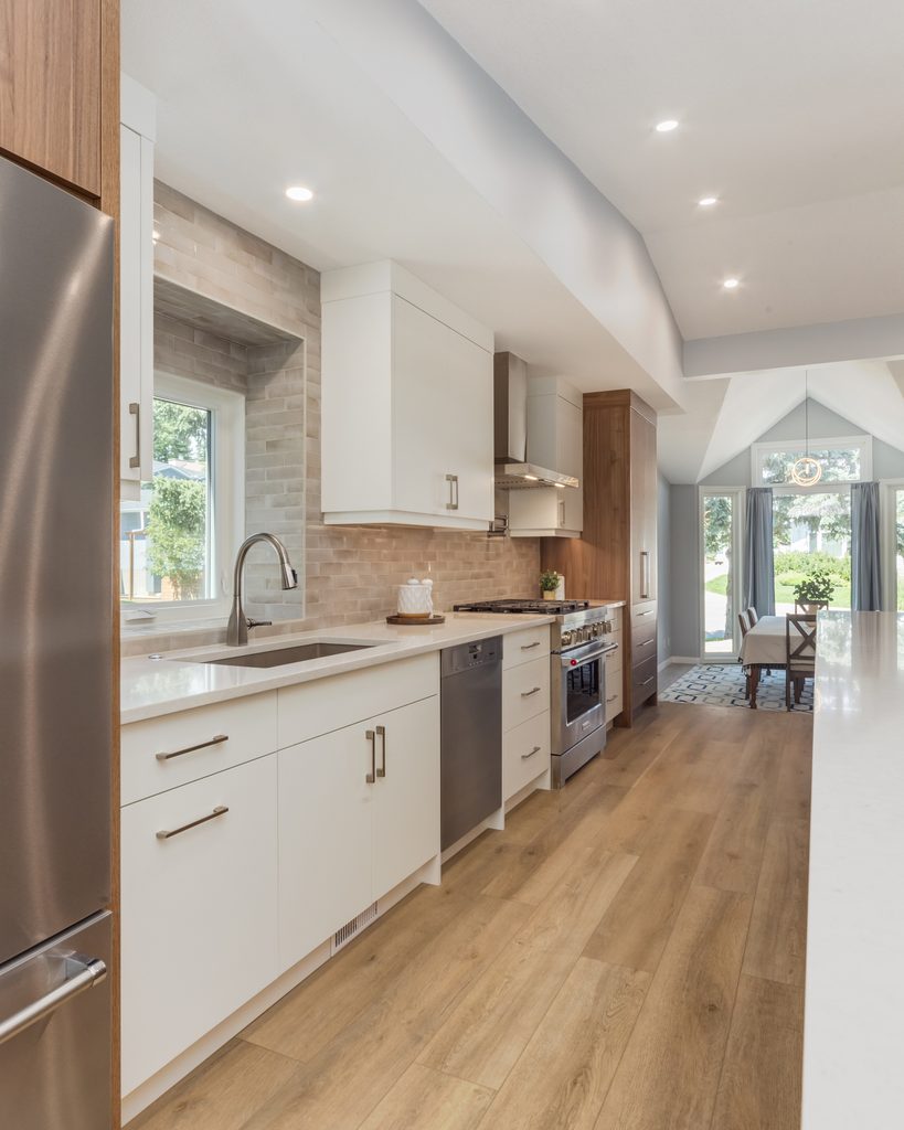Modern kitchen with white cabinets, stainless steel appliances, light wood flooring, a large window over the sink, and an open layout leading to a bright dining area with patio doors.