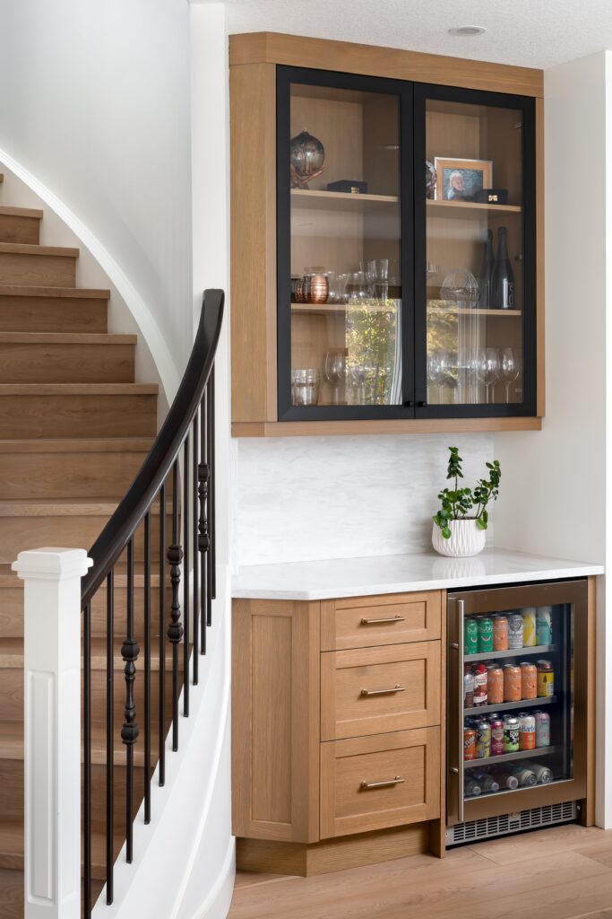 A small home beverage station with wood cabinets, a glass-front upper cabinet, a potted plant, and a mini fridge stocked with drinks, situated next to a staircase with a black railing.