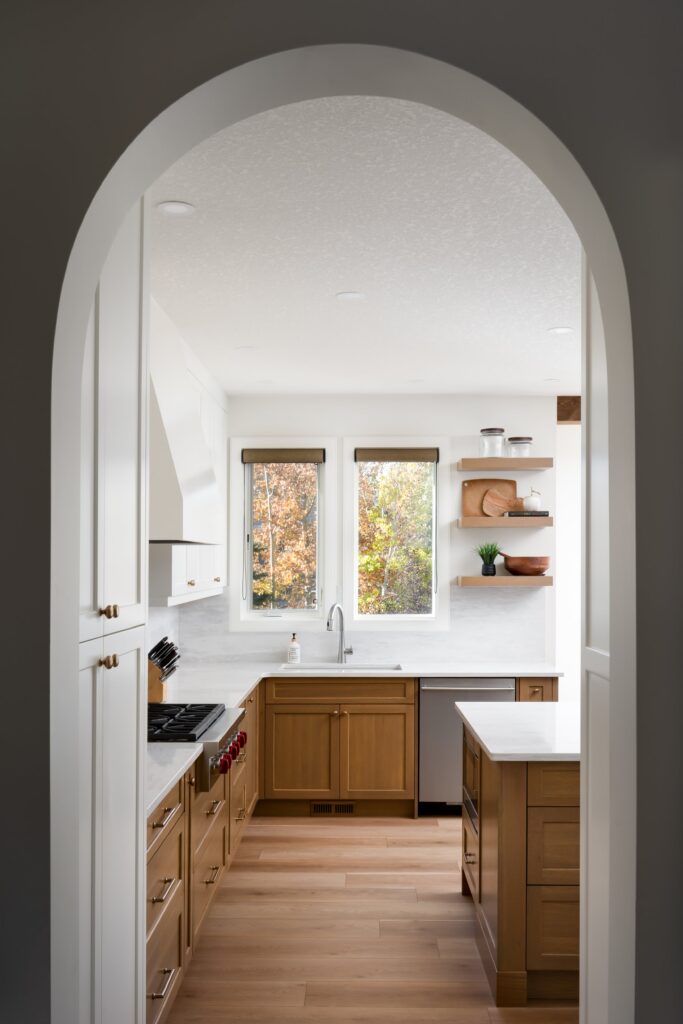 Modern kitchen with light wood cabinets, white countertops, and an island, viewed through an arched doorway. Two windows above the sink let in natural light. Open shelves display dishes and decor items.