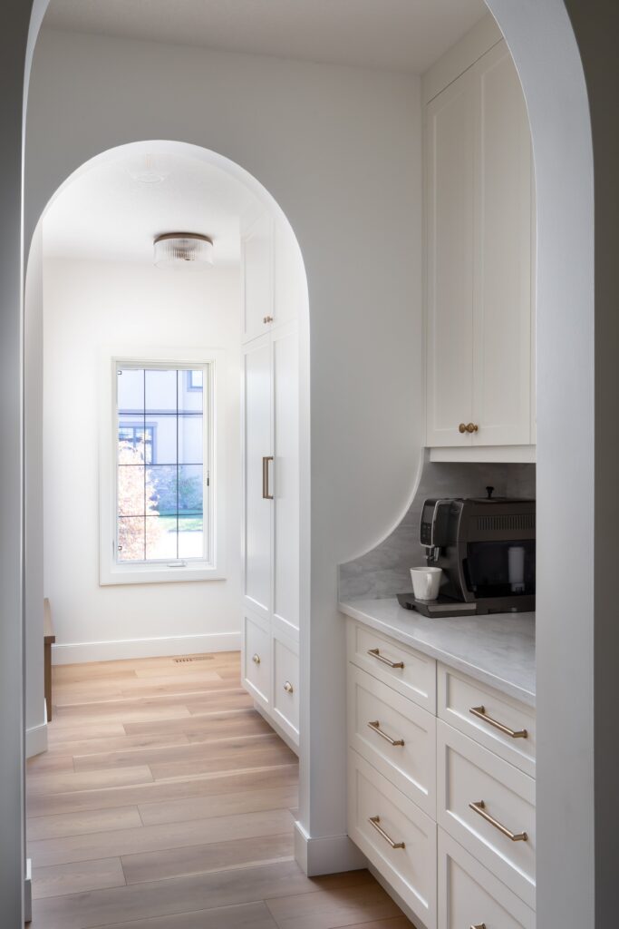 A bright, modern kitchen with white cabinets, gold handles, light wood floors, a coffee machine on the counter, and an arched doorway leading to a window with natural light.