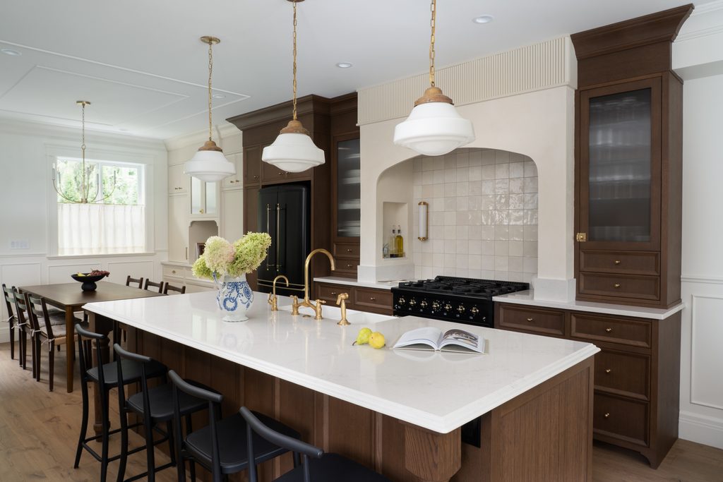 A modern kitchen features a large island with a white countertop, black chairs, pendant lights, wood cabinets, a black stove, and a vase with hydrangeas. Lemons and an open book rest on the island.