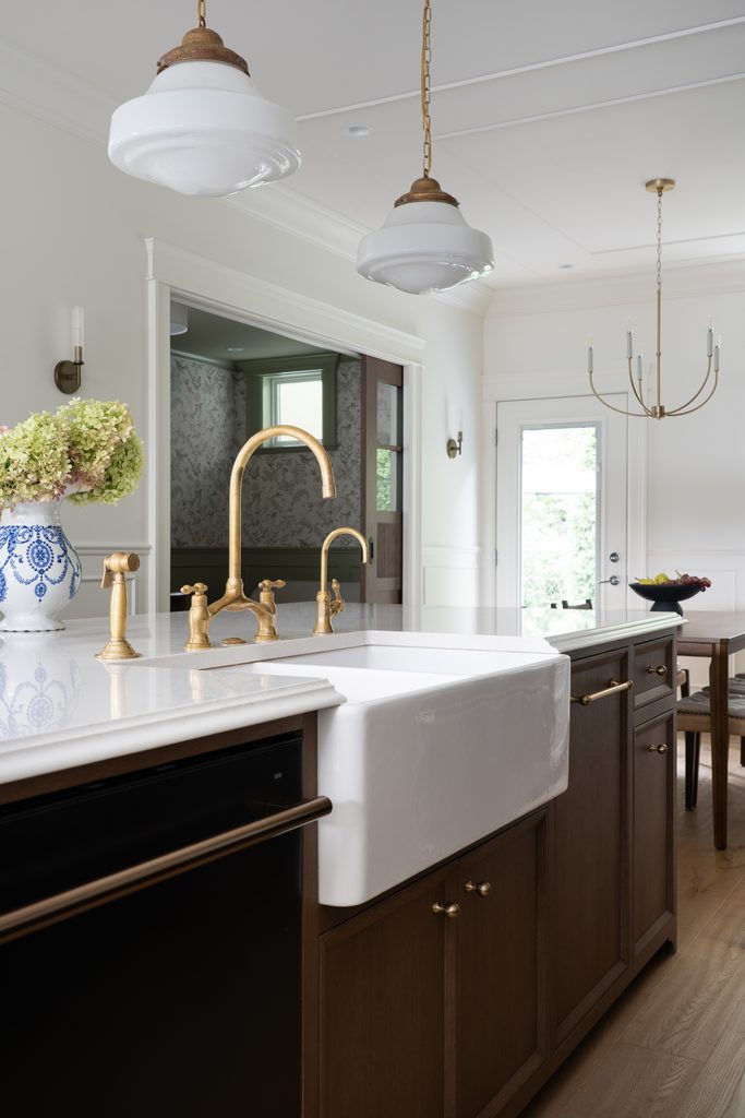 Modern kitchen with a white farmhouse sink, brass faucet, white countertops, dark wood cabinets, pendant lights, chandelier, and a blue-and-white vase with flowers. Dining area and door are visible in the background.