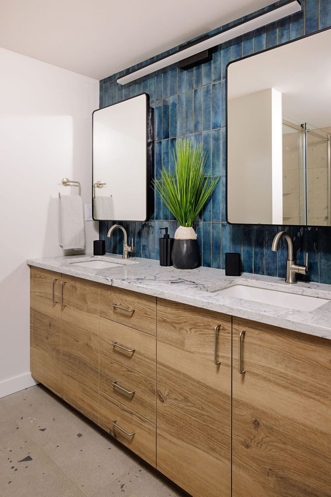 Modern bathroom with double sinks, wood cabinetry, marble countertop, two mirrors, blue tile backsplash, wall-mounted faucets, and a vase with green foliage on the counter. Towels are hanging on the wall.