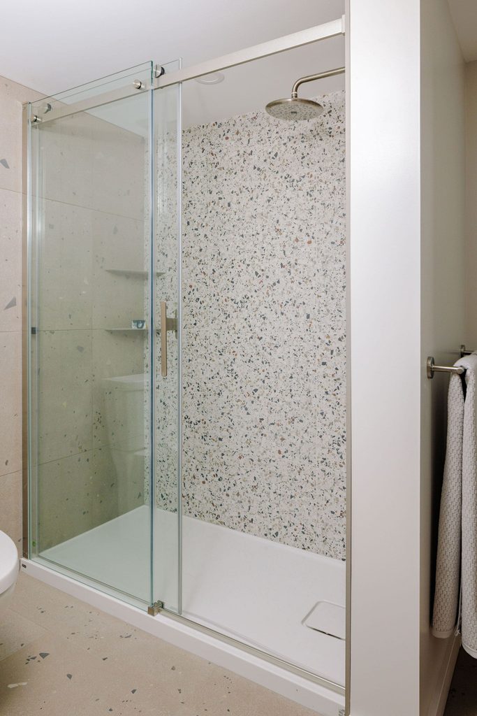 Modern bathroom with a glass shower enclosure, terrazzo-style wall tiles, a silver rain showerhead, and a towel hanging on a rack outside the shower.