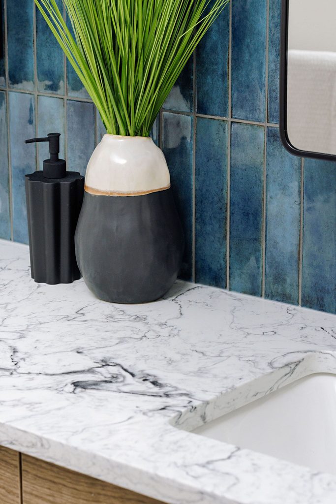 A marble countertop with a black soap dispenser, a ceramic vase holding green grass, and blue tile backsplash is shown next to a white sink and a mirror.