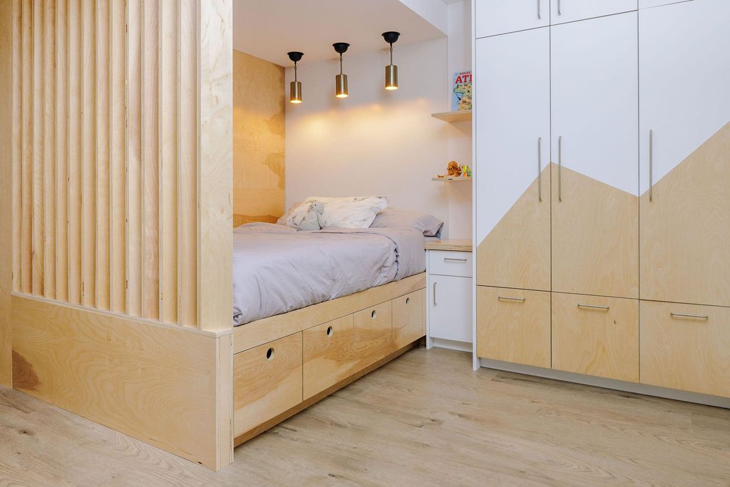 Modern bedroom with a built-in light wood bed, storage drawers underneath, wall-mounted lights above, and a white closet with geometric designs. Light wood flooring and minimal decor create a clean, cozy look.