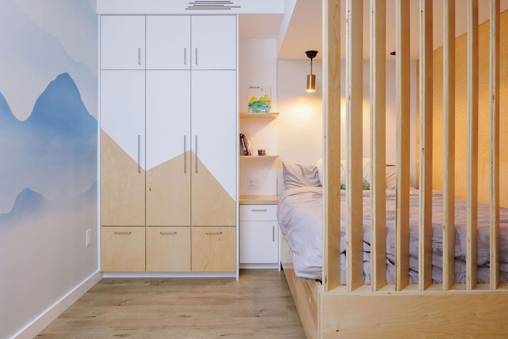 A modern bedroom with a wooden floor, a built-in wardrobe with a mountain design, a cozy bed behind wooden slats, and a soft blue mountain mural on the wall. Theres a small shelf and light by the bed.