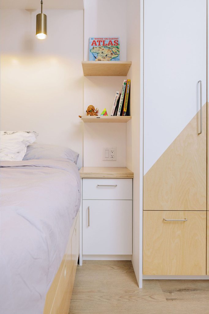 A tidy bedroom corner with a bed, built-in shelves holding books and toys, a cabinet, and a soft hanging light above. The decor is minimalist with light wood and white finishes.