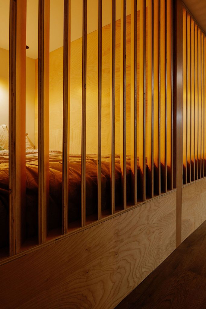 A cozy bedroom with warm lighting, viewed through vertical wooden slats that partially enclose the bed, creating a sense of privacy and a modern, minimalist design.