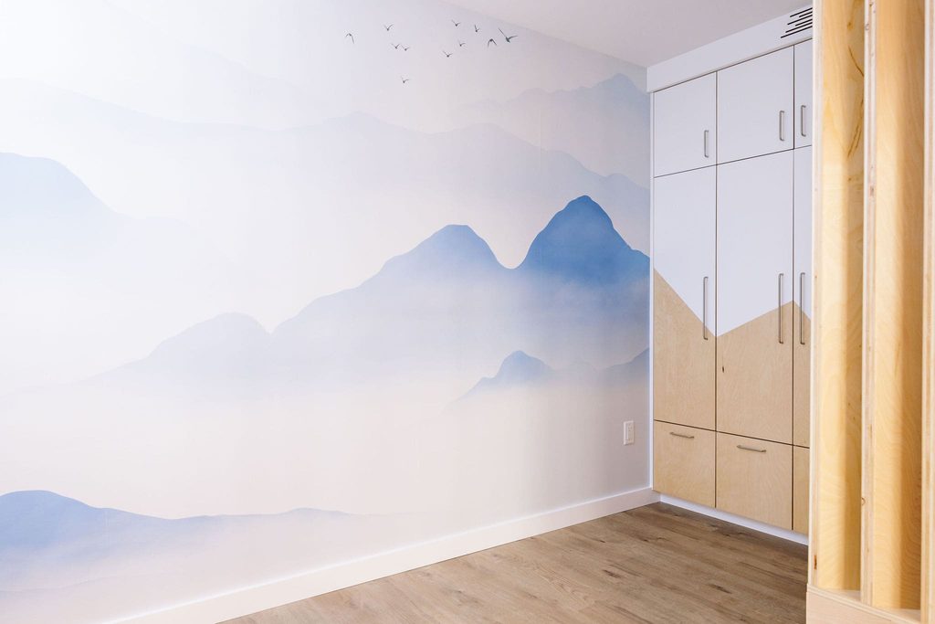 A minimalist room with a wooden floor, a built-in cabinet, and a wall mural depicting soft blue mountains and birds in the sky.