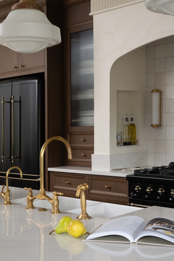 Modern kitchen with gold fixtures, white countertop, dark wood cabinets, black refrigerator, open cookbook, two pears, and olive oil bottles near a white tile backsplash and gas stove.