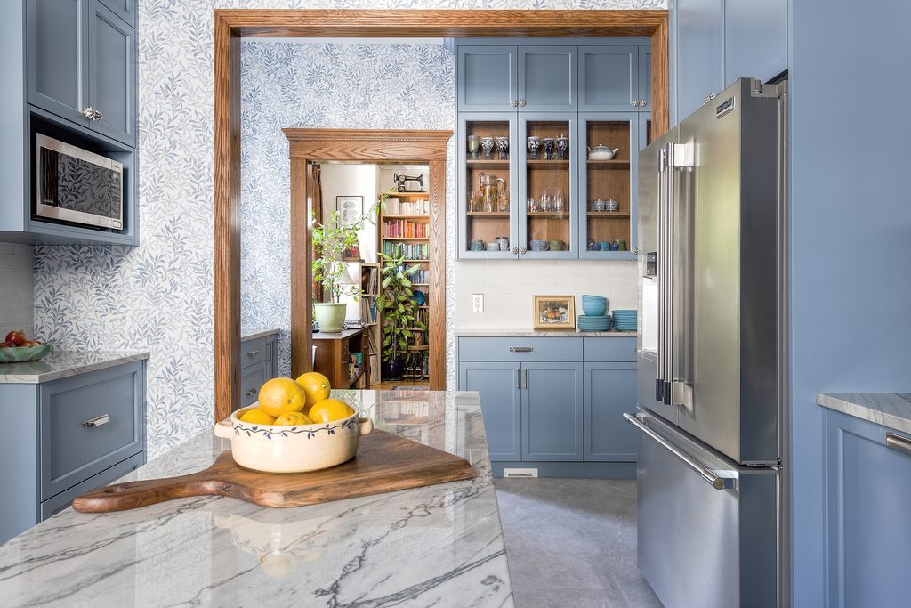 A modern blue kitchen with marble countertops, stainless steel fridge, glass cabinet doors, a bowl of lemons on a wooden board, and a view into a cozy, plant-filled room with bookshelves.