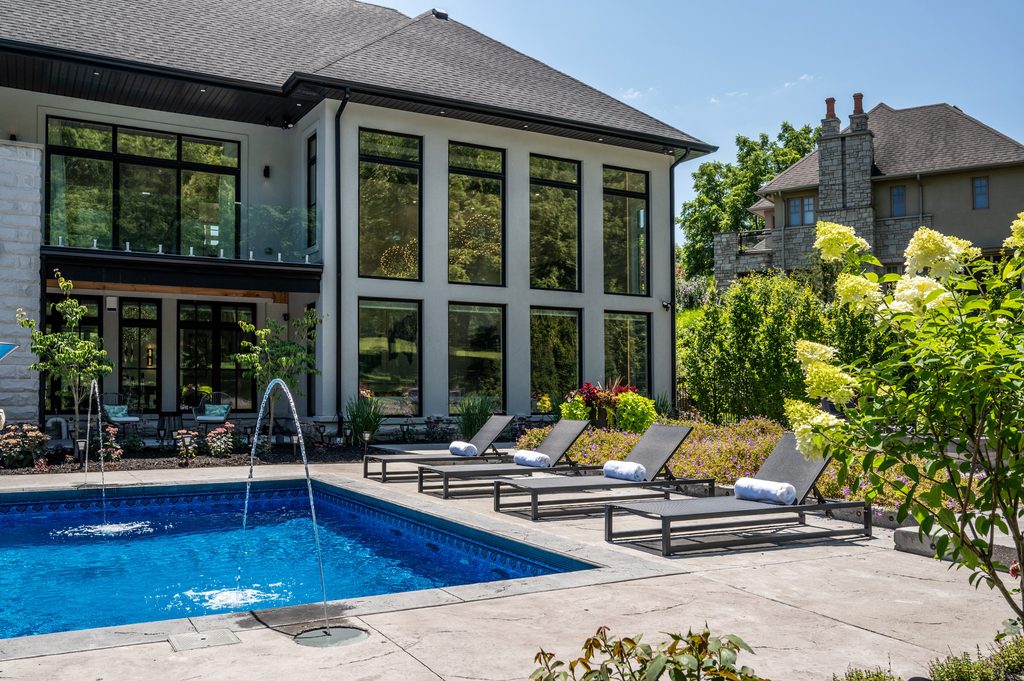 Modern backyard with a blue swimming pool, four lounge chairs with towels, blooming flowers, and a two-story house with large windows and a balcony in the background.