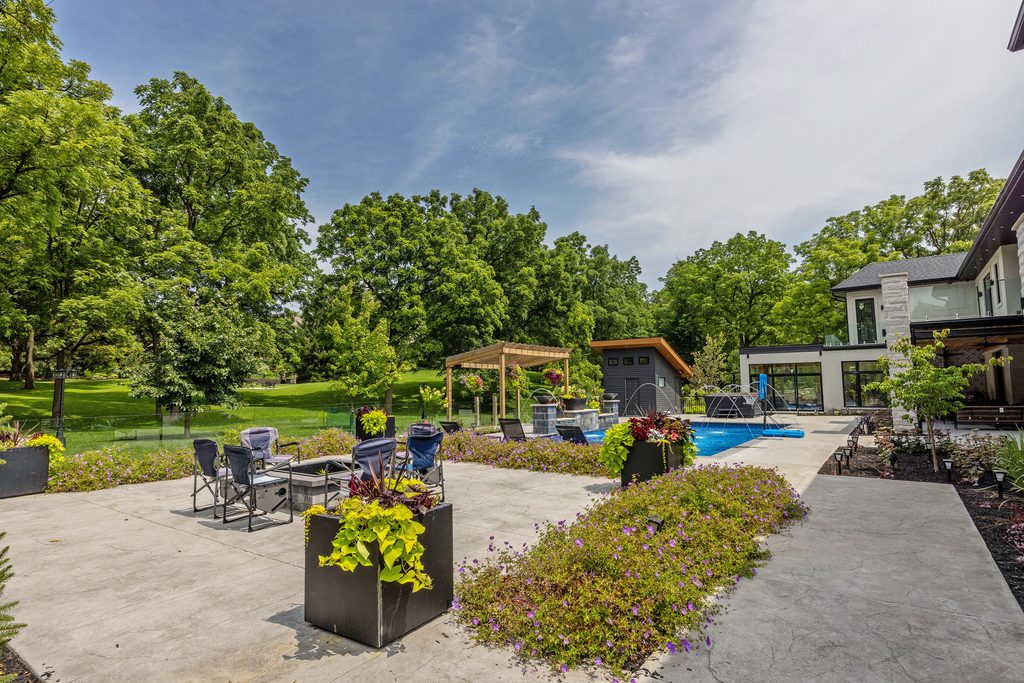 A spacious backyard features a patio with chairs, lush planters, a swimming pool, and a pergola. Green lawn and trees surround the area, with a modern house on the right under a partly cloudy sky.