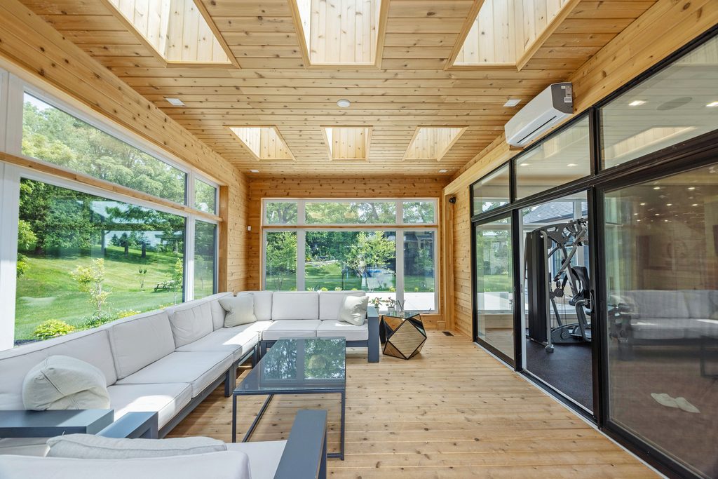 A bright sunroom with wooden walls and ceiling, skylights, large windows, a white sectional sofa, glass coffee table, and garden view outside.