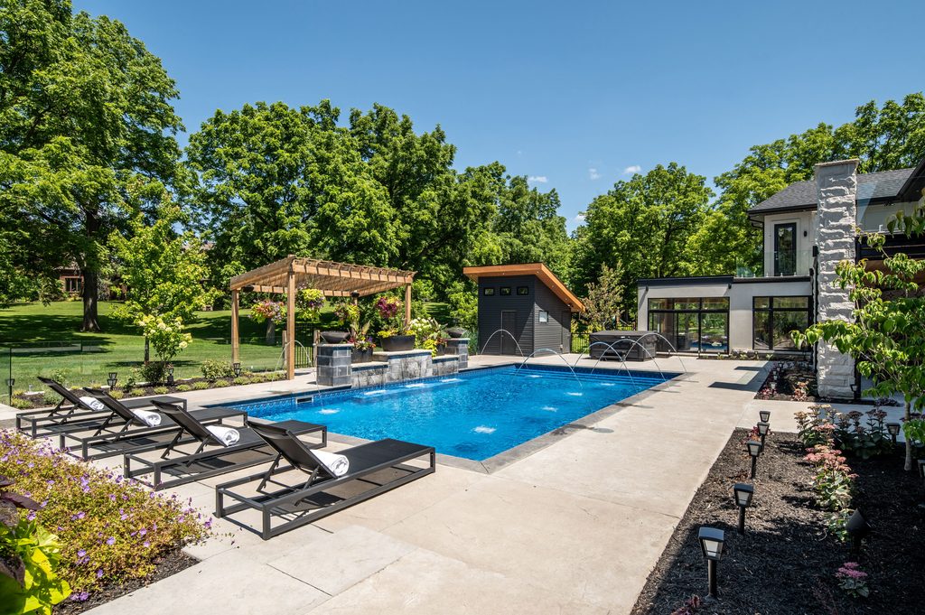 Modern backyard with a rectangular pool, four black lounge chairs, a pergola, and landscaped greenery. A gray pool house and a contemporary home are visible in the background under a clear blue sky.