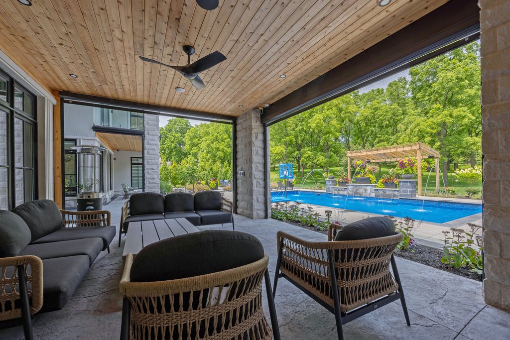 Covered patio with wicker furniture overlooks a backyard swimming pool, surrounded by greenery and a wooden pergola. Ceiling fan and recessed lighting are visible; large glass doors open to the outdoor space.