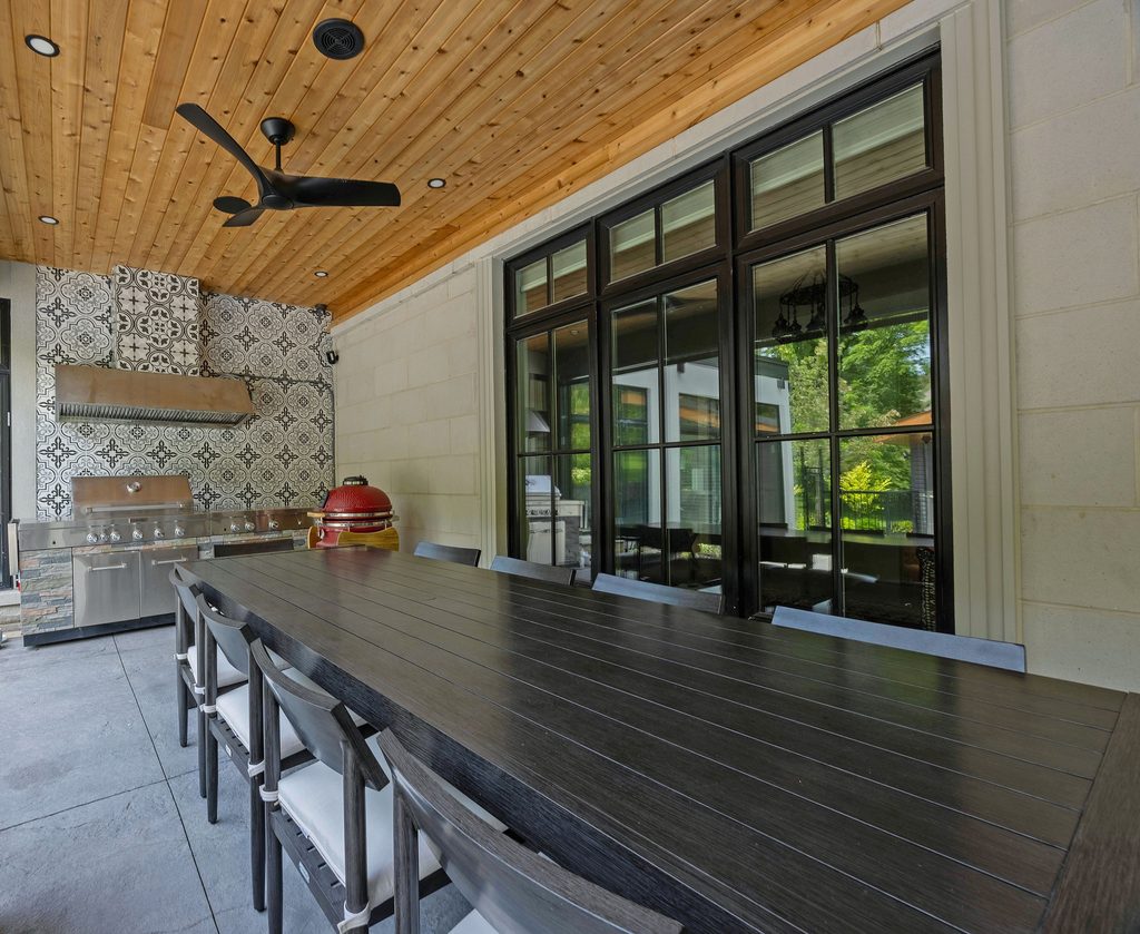 Covered outdoor dining area with a long dark table, modern black chairs, a ceiling fan, and a grill station featuring patterned tiles and a red ceramic grill. Large windows reflect greenery outside.