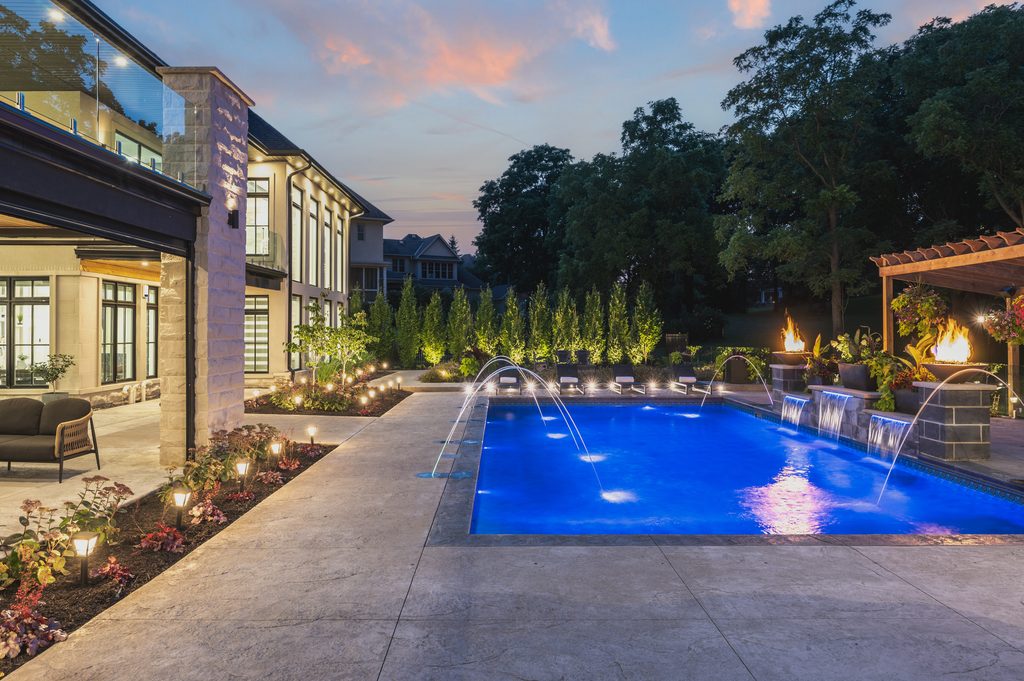A modern backyard features a lit swimming pool with water fountains, surrounded by a patio, seating areas, fire pits, lush greenery, and soft evening lighting.