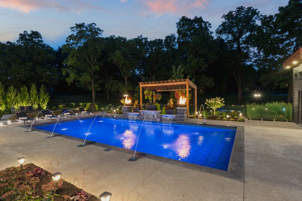 A lit swimming pool at dusk with water fountains, fire features, a pergola, and lush landscaping. Trees and pathway lights surround the modern outdoor area.