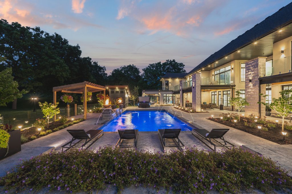A modern luxury home at dusk features a glowing blue swimming pool, lounge chairs, pergola, and landscaped yard with trees and flowers, all illuminated by garden lights.